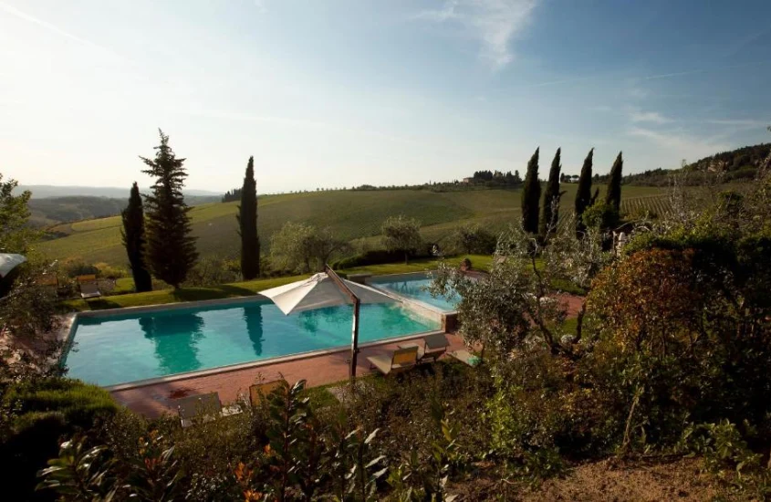 Piscina rodeada por jardins exuberantes, com vista para as colinas e vinhedos típicos da Toscana, em um cenário tranquilo e ensolarado.