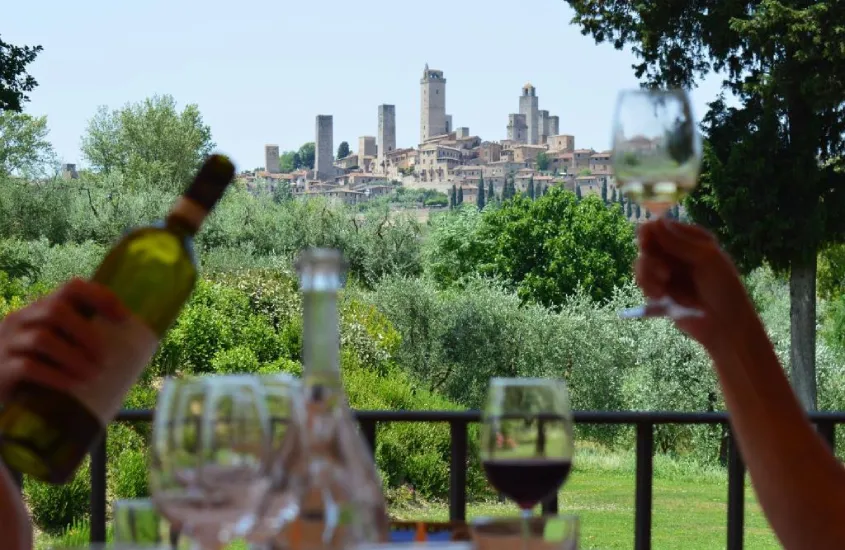 Pessoas brindando com taças de vinho em um terraço com vista para San Gimignano, com suas torres icônicas e vegetação ao redor.