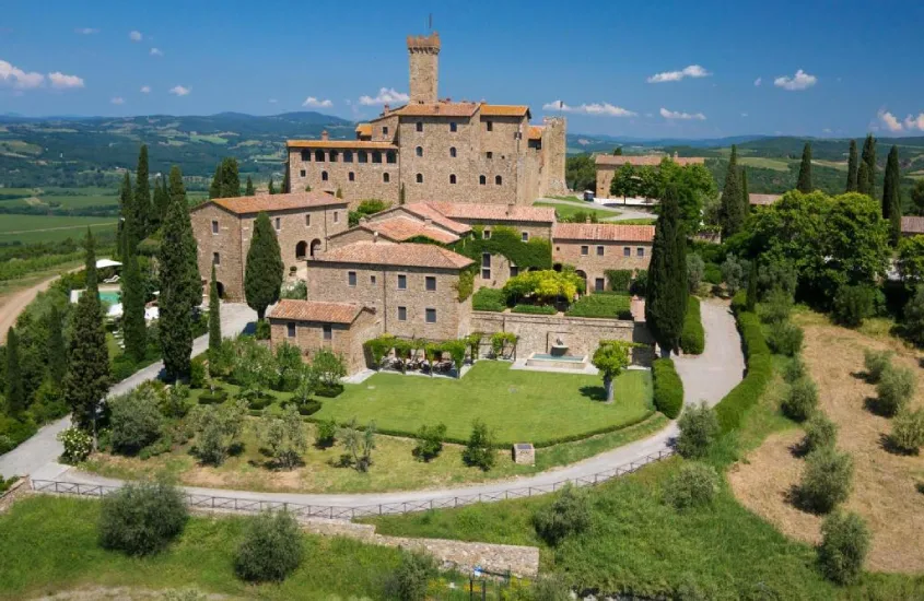 Vista aérea de um imponente castelo medieval cercado por jardins bem cuidados, ciprestes e vinhedos que se estendem ao horizonte.