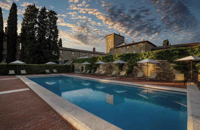 Piscina com borda em estilo clássico rodeada por espreguiçadeiras e guarda-sóis, com uma antiga construção de pedra e árvores altas compondo o cenário.