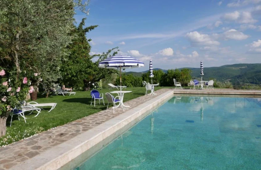 Piscina ladeada por gramado e árvores floridas, com cadeiras e guarda-sóis decorando o espaço em meio às colinas verdes da Toscana.