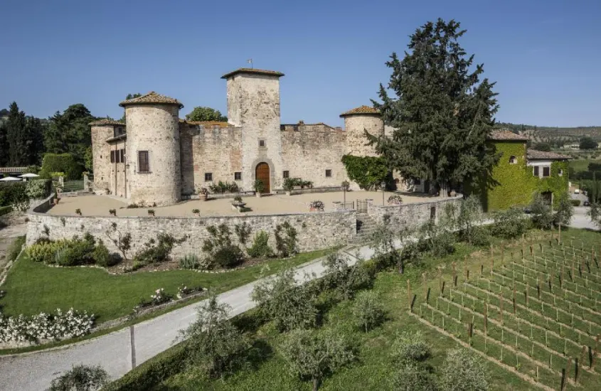 Castelo histórico com torres de pedra e fachada coberta de hera, rodeado por vinhedos, jardins simétricos e estradas de cascalho.