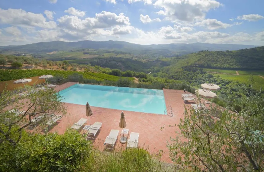 Piscina com borda infinita oferecendo uma vista panorâmica para os vinhedos e vales da Toscana, sob um céu claro e ensolarado.