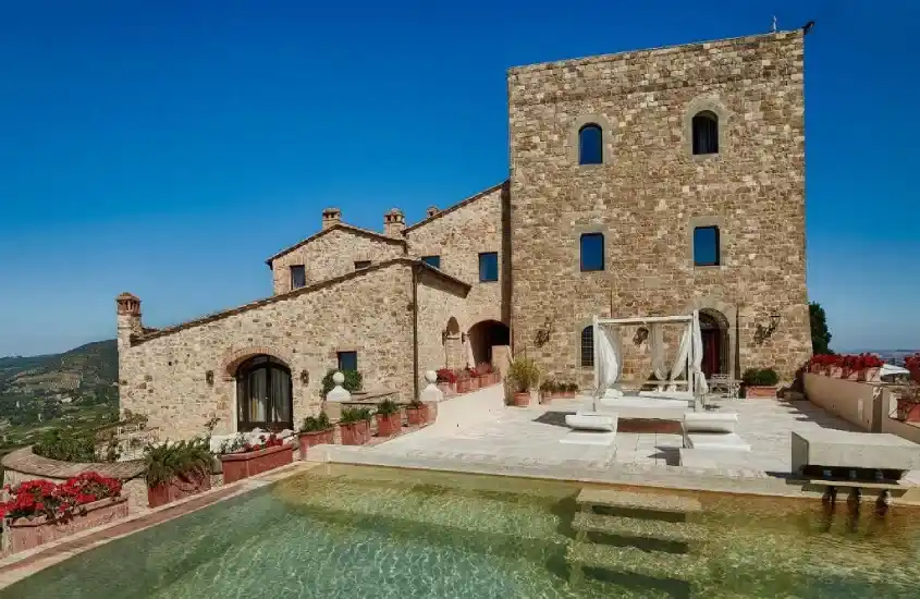 Construção de pedra clara com detalhes rústicos, rodeada por uma piscina termal com vista para as paisagens da Toscana.