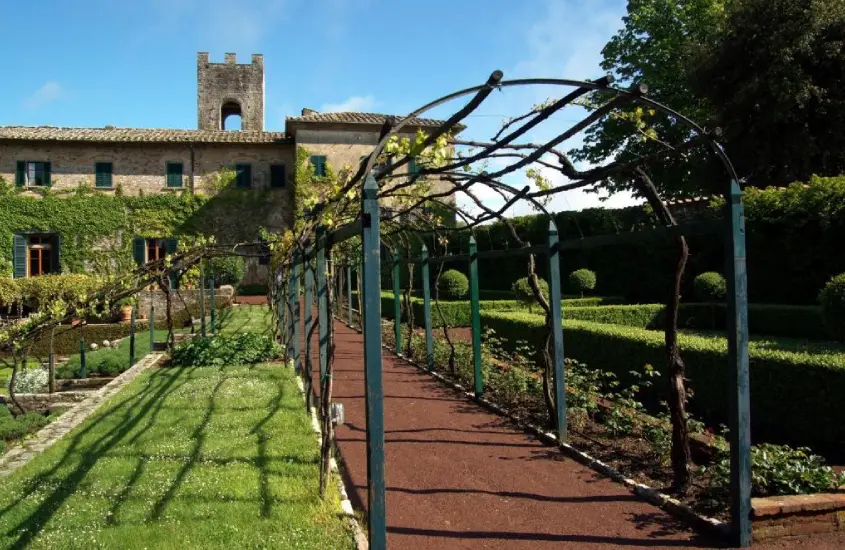 Caminho arborizado com pérgula de videiras conduzindo a uma construção histórica com jardins perfeitamente cuidados.