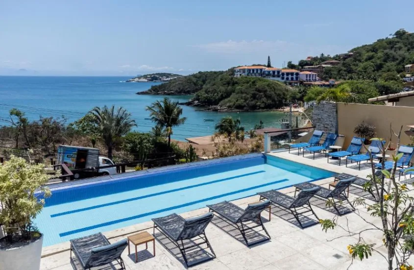 Em um dia de sol, área de lazer de hotel com recreação infantil em Búzios com piscina, espreguiçadeiras, árvores ao redor e praia na frente