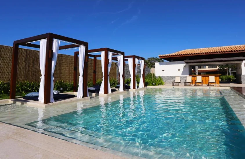 Em um dia de sol, área de lazer de hotel com piscina, tendas com camas, espreguiçadeiras e plantas ao redor