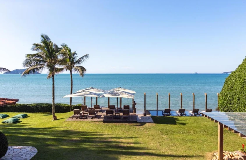 Em um dia de sol, área gramada de hotel com camas, guarda-sóis, árvores ao redor e praia na frente