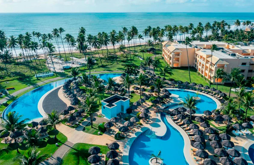 Em um dia de sol, vista aérea de hotel com piscinas, espreguiçadeiras, guarda-sóis, árvores ao redor e praia do lado