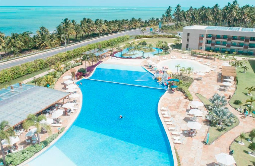 Num dia ensolarado, vista aérea de um hotel com piscina grande, espreguiçadeiras, praia do lado e árvores ao redor