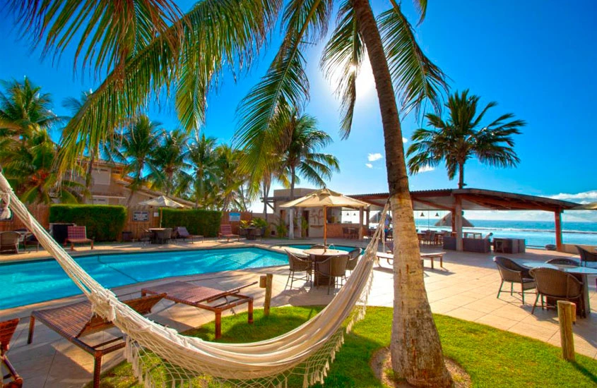 Uma rede de descanso amarrada entre duas palmeiras ao lado de uma piscina em um ambiente tropical. O sol brilha intensamente, destacando o azul do céu e o mar ao fundo