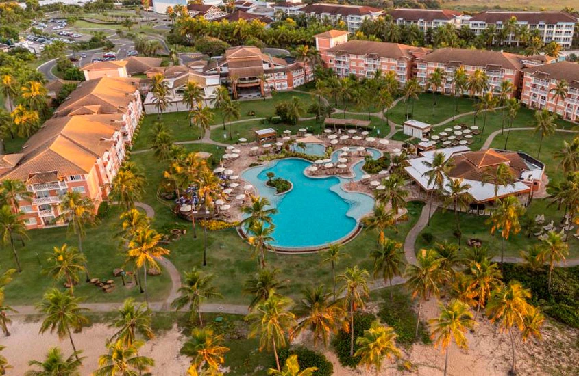 Vista panorâmica de um resort com prédios de telhado laranja, uma grande piscina em formato orgânico cercada por gramados bem cuidados e muitas palmeiras. Ao fundo, dá para ver o estacionamento e outras estruturas do local