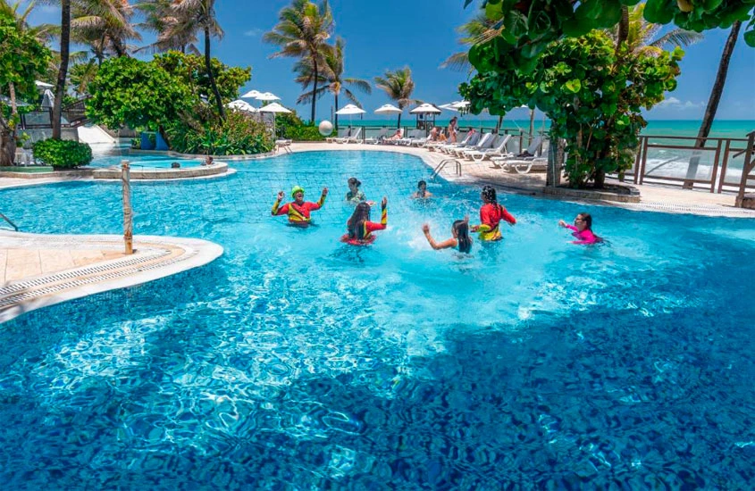 Pessoas jogando vôlei aquático em uma piscina cercada por coqueiros, guarda-sóis brancos e um ambiente tropical. O céu azul brilhante realça a atmosfera de lazer e diversão.