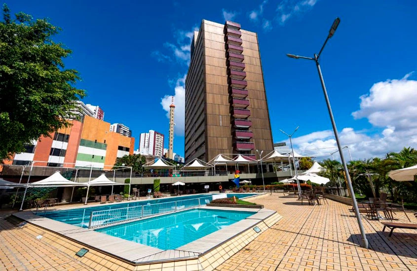 Em um dia de sol, área de lazer de um hotel com recreação infantil no nordeste com piscinas, espreguiçadeiras, árvores ao redor e guarda-sóis