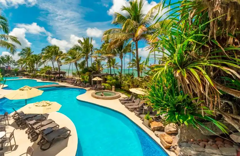 Em um dia de sol, área de lazer de um hotel com recreação infantil no nordeste com piscina, espreguiçadeiras, guarda-sóis e árvores ao redor