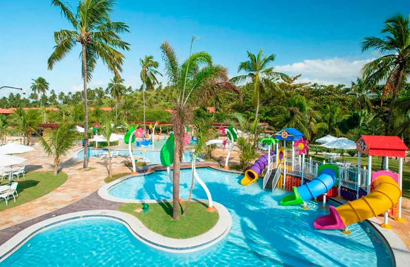 Em um dia ensolarado, vista de uma área de lazer com piscinas, espreguiçadeiras, guarda-sóis, brinquedos de água e árvores ao redor