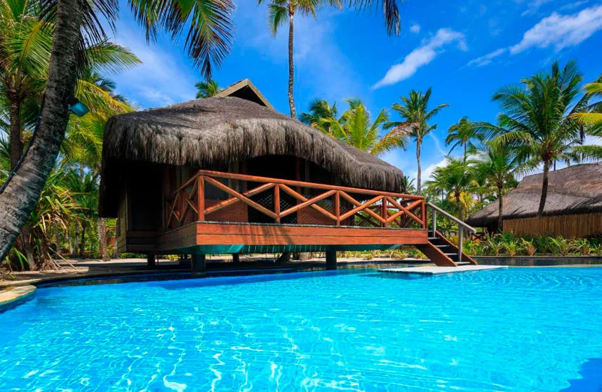 Em um dia de sol, área de piscina de um hotel com recreação infantil no nordeste com tenda de palha e árvores ao redor