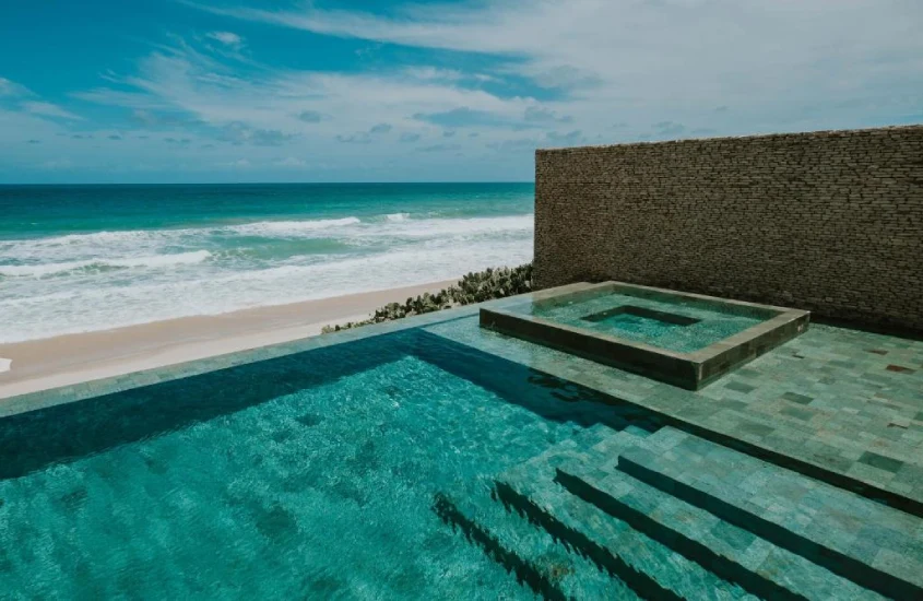 Piscina com borda infinita que se funde à vista do mar, com um design minimalista cercado por uma parede de pedra, criando um ambiente de luxo à beira da praia.