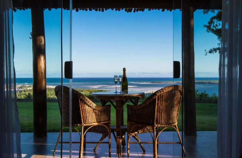 Mesa com cadeiras de vime em uma varanda com vista para o mar, decorada com taças e uma garrafa de espumante, criando um ambiente romântico e tranquilo.