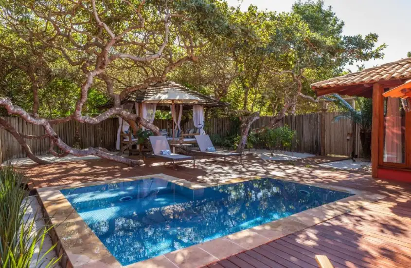 Área com piscina cercada por árvores retorcidas, um gazebo com espreguiçadeiras e um deck de madeira, criando um ambiente privativo e tranquilo.