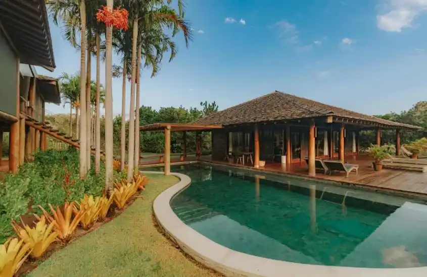 Piscina ao ar livre com bordas sinuosas, rodeada por palmeiras e jardins bem cuidados, complementada por uma ampla estrutura em madeira com varanda coberta.