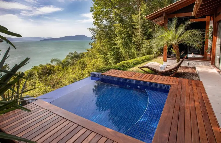 Piscina com revestimento em mosaico azul e borda de madeira, rodeada por vegetação exuberante, com vista para o mar ao longe.