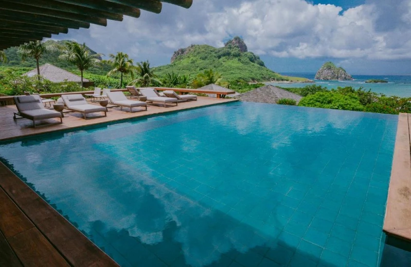 Piscina com borda infinita e espreguiçadeiras ao redor, destacando uma vista deslumbrante de Fernando de Noronha, com colinas verdes ao fundo.