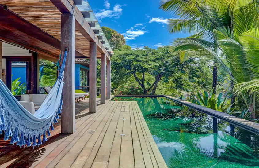 Piscina de borda infinita cercada por um amplo deck de madeira, rodeada por vegetação tropical, com um visual sereno e exuberante.