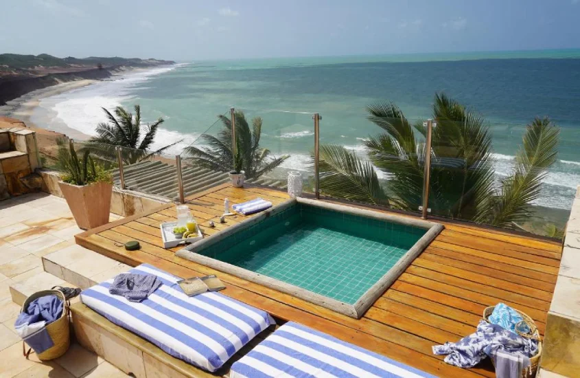 Jacuzzi com borda de madeira e espreguiçadeiras ao lado, situada em um terraço com vista panorâmica para o mar e falésias, cercada por palmeiras.