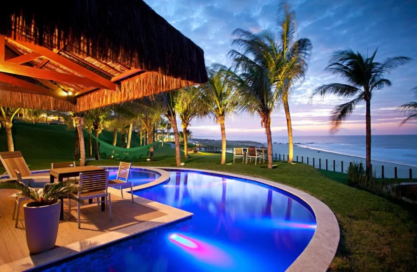 Piscina iluminada à noite com luzes coloridas, cercada por coqueiros e áreas de descanso, com vista para o mar em um cenário tropical.