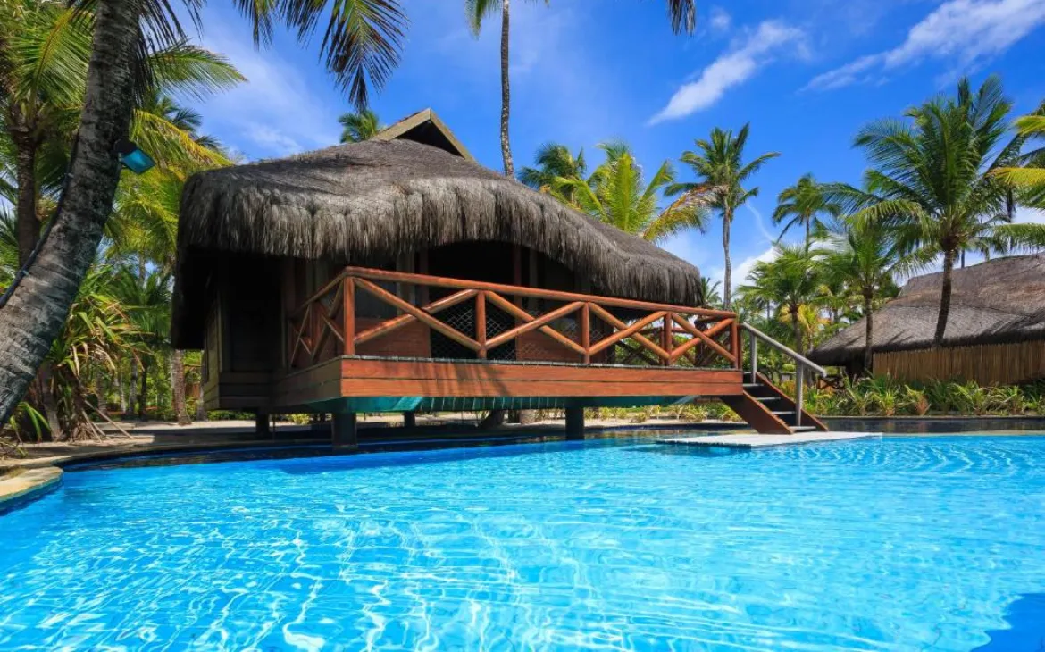 Bangalô de madeira com telhado de palha, cercado por palmeiras e localizado à beira de uma piscina azul cristalina em hotel estilo Maldivas no Brasil