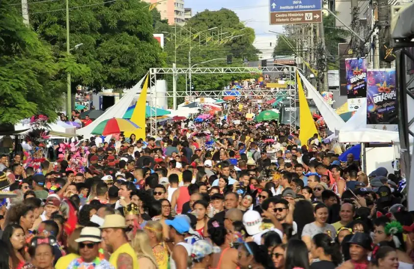 Ruas lotadas de foliões em um bloco de Carnaval ao ar livre, com tendas coloridas, guarda-sóis e decoração temática, em um clima de festa e descontração.