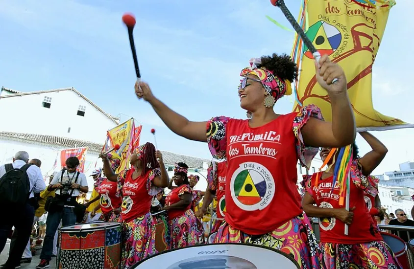 Mulheres vestidas com trajes coloridos e tocando tambores em uma apresentação cultural ao ar livre, com bandeiras ao fundo e muita alegria.