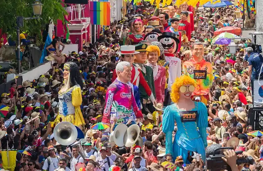 Bonecos gigantes desfilando pelas ruas lotadas de Olinda, rodeados por foliões em clima de festa, com decoração colorida e casarios históricos ao fundo.