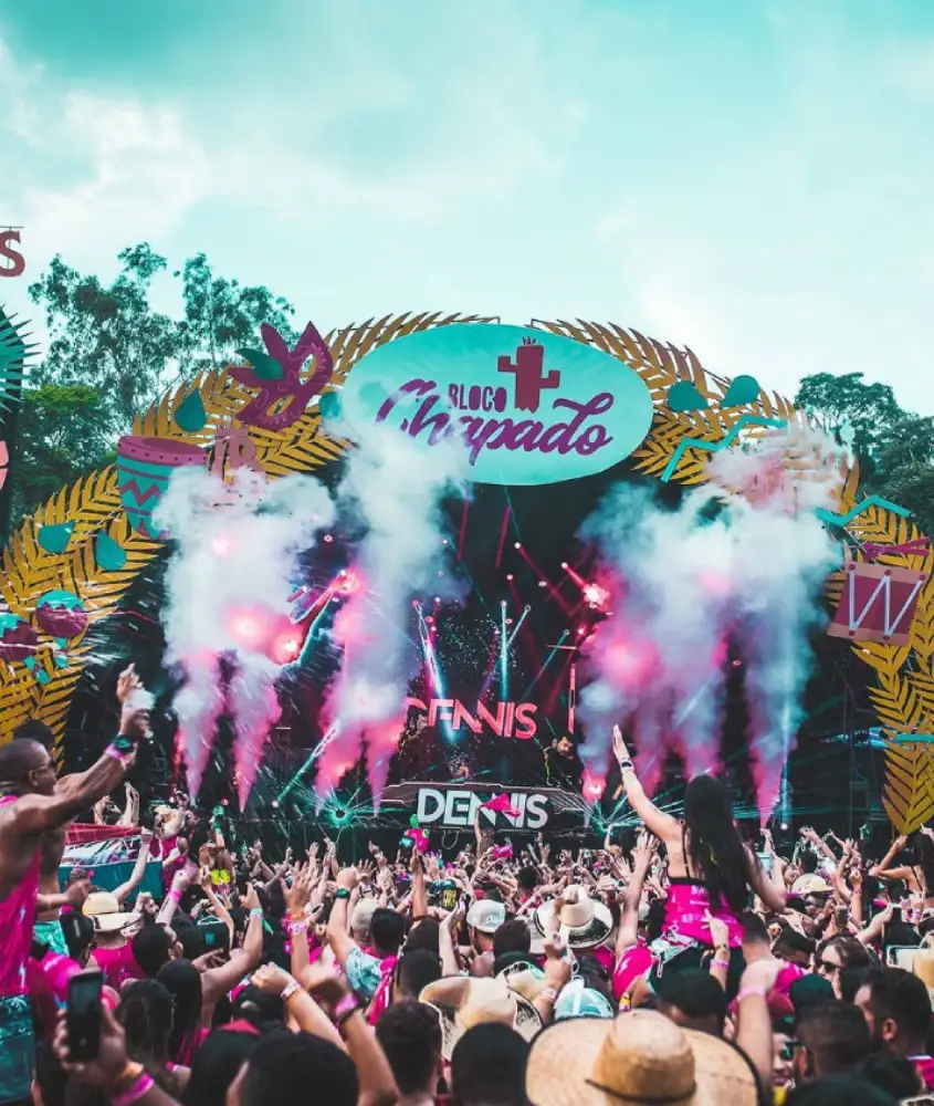 Palco de um bloco de Carnaval moderno, cercado por foliões vestidos de rosa, luzes coloridas e fumaça, com um cenário tropical ao fundo.