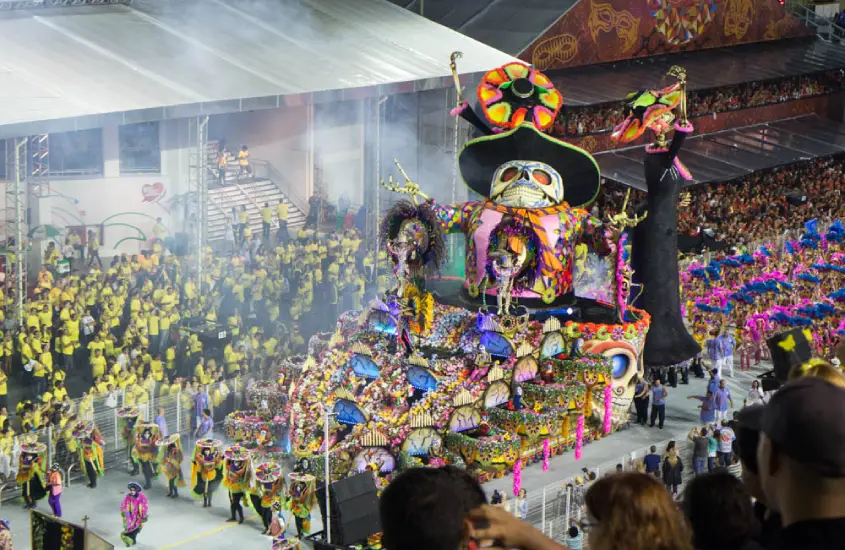 Alegoria colorida em um desfile de escola de samba, com detalhes temáticos e figurinos vibrantes, em um sambódromo repleto de espectadores.