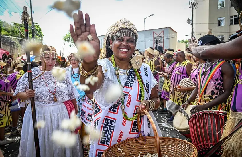 Mulher sorridente vestida com trajes brancos e colares coloridos, distribuindo grãos de pipoca em uma celebração tradicional de matriz africana, cercada por tambores e participantes caracterizados.