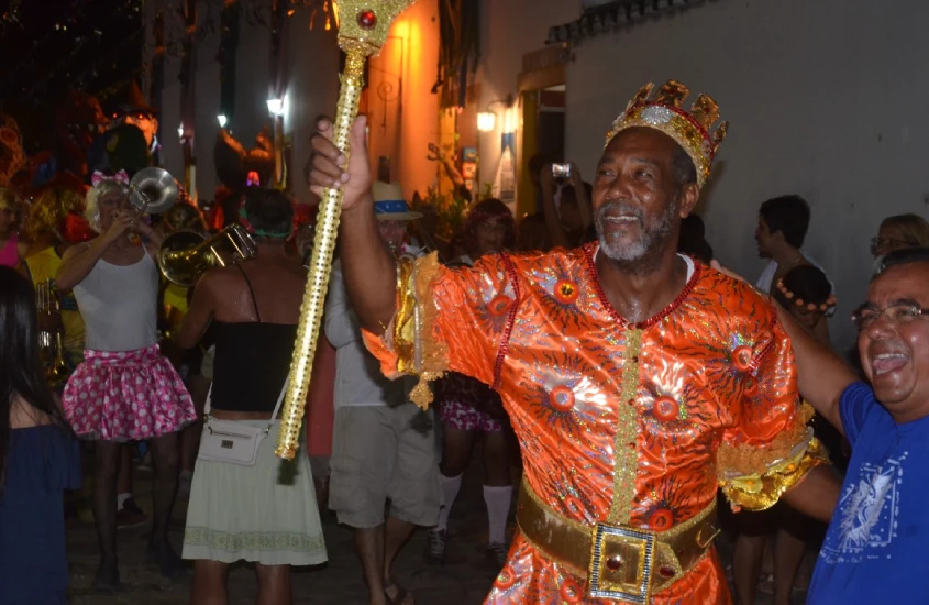 Homem em traje laranja brilhante e coroa dourada, conduzindo um desfile noturno de Carnaval com músicos e foliões fantasiados, em um ambiente festivo e iluminado.