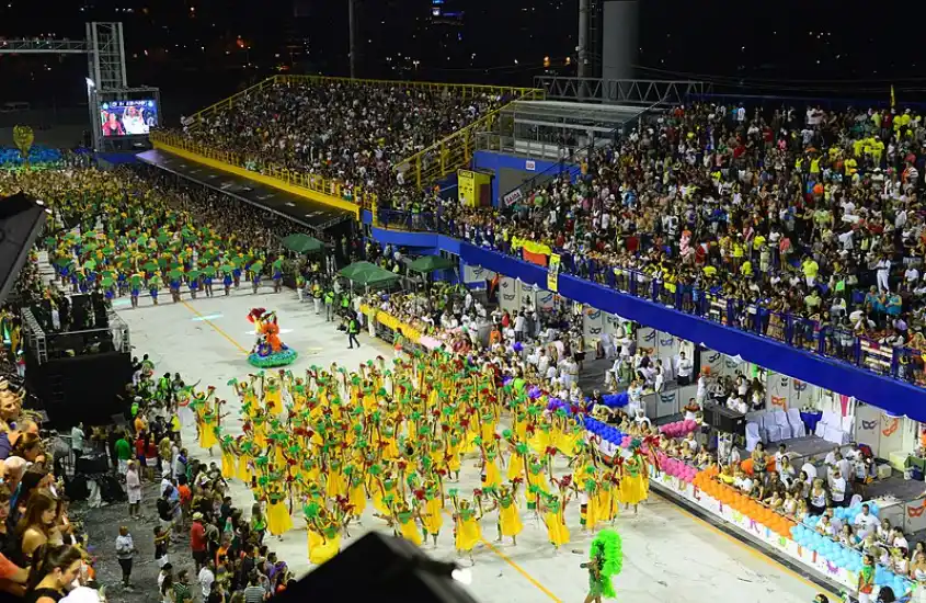 Desfile noturno de escola de samba no sambódromo, com alas coloridas e arquibancadas lotadas de espectadores, em uma celebração vibrante e cheia de energia.