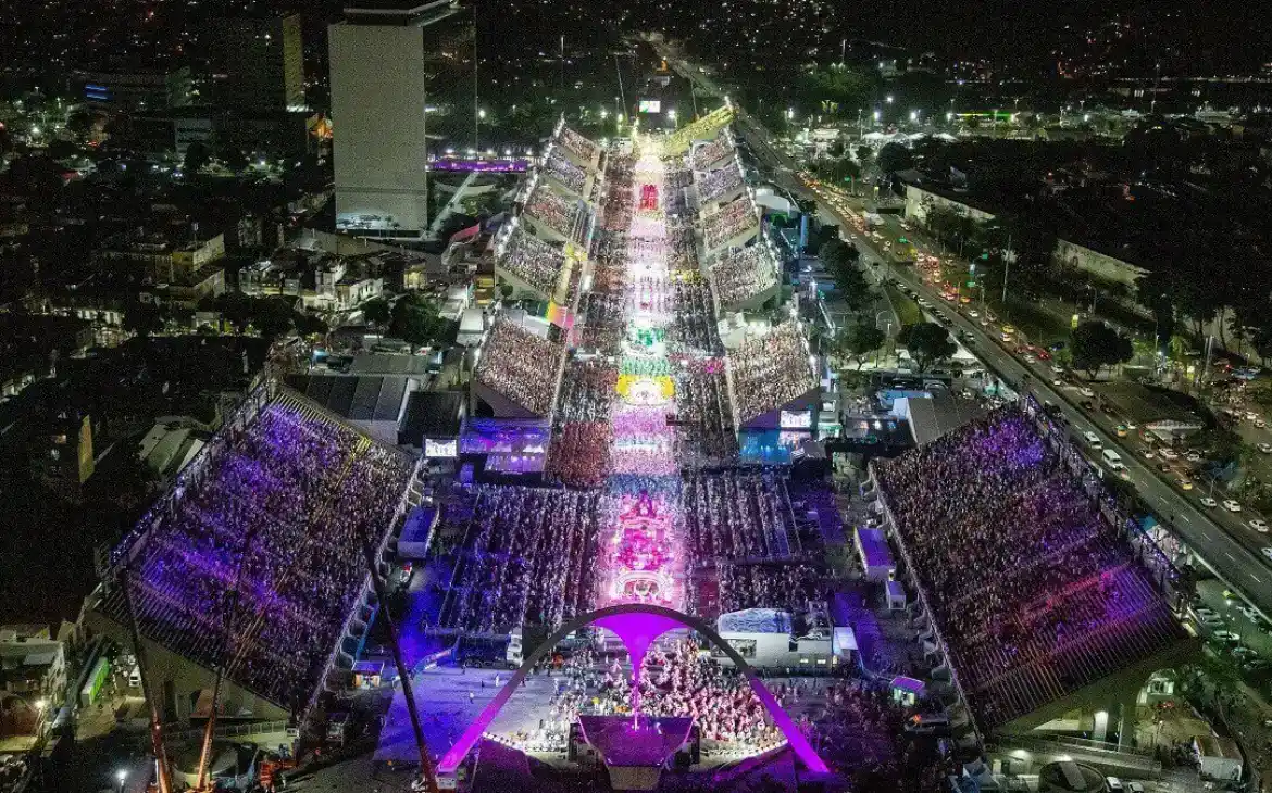 Vista aérea do sambódromo iluminado durante um desfile noturno de Carnaval, com arquibancadas lotadas e alegorias desfilando pela avenida central