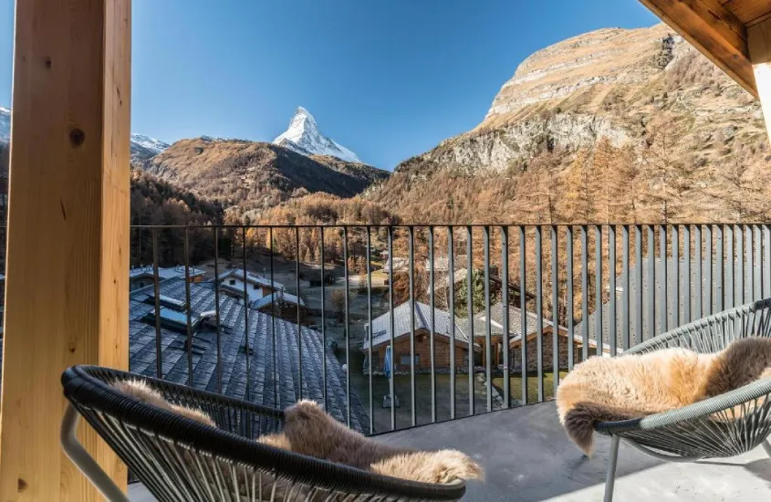 Varanda com duas cadeiras forradas e uma vista ampla das montanhas ao fundo, destacando o icônico Matterhorn cercado por árvores e casas.