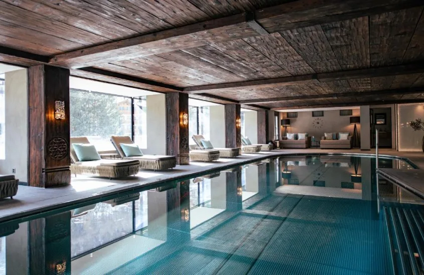 Piscina interna com design aconchegante, cercada por cadeiras de descanso, detalhes em madeira e amplas janelas que permitem entrada de luz natural.