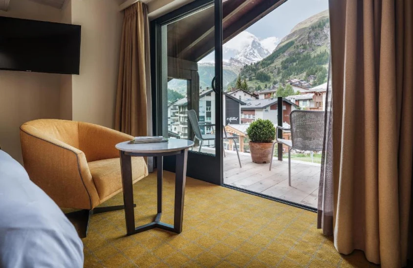 Quarto de hotel com uma poltrona amarela, mesa lateral e vista para uma varanda com o Matterhorn ao fundo, emoldurada por casas e montanhas verdes.