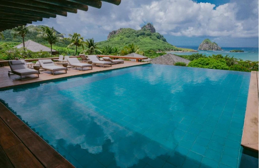 Piscina de borda infinita cercada por espreguiçadeiras brancas com vista para o mar e montanhas cobertas de vegetação ao fundo.