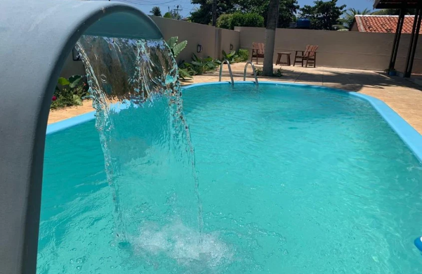 Piscina com cascata ao ar livre, em ambiente cercado por parede e algumas plantas ao redor, sob um céu claro.