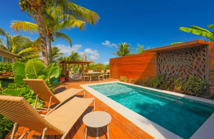 Área de piscina da Pousada Atobá com espreguiçadeiras de vime sob a sombra de coqueiros e plantas tropicais