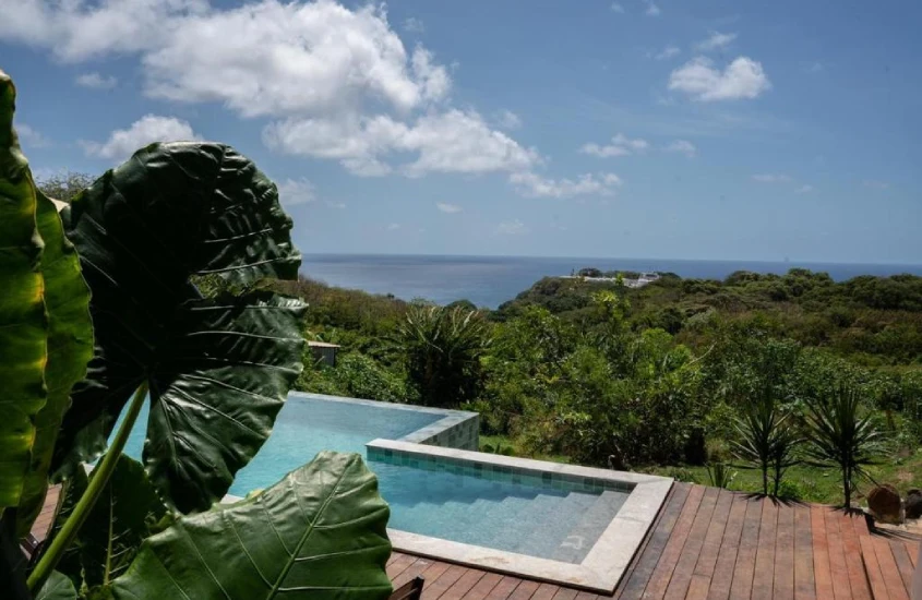 Vista da piscina com borda infinita da Miragem Noronha, com folhagens tropicais em primeiro plano e o oceano e montanhas ao fundo.