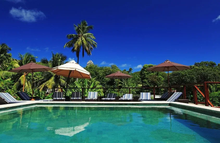 Piscina ao ar livre com guarda-sóis e espreguiçadeiras listradas, rodeada por palmeiras e vegetação sob um céu azul.