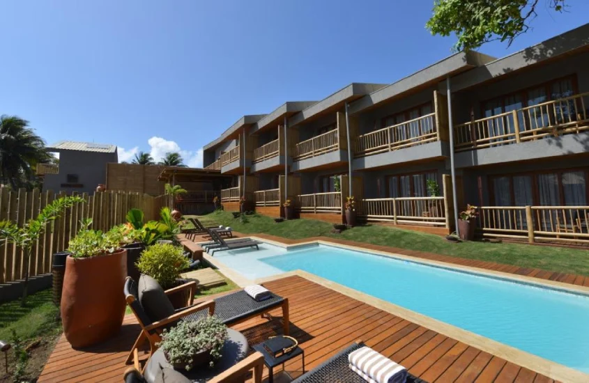 Piscina retangular rodeada por deck de madeira e cercada por vegetação tropical, com cadeiras dispostas ao lado de um prédio de dois andares com varandas.