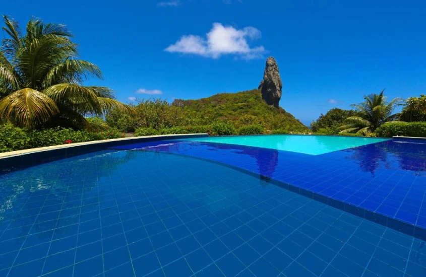 Piscina de borda infinita com vista para o icônico Morro do Pico em Fernando de Noronha, cercada por vegetação e céu azul.